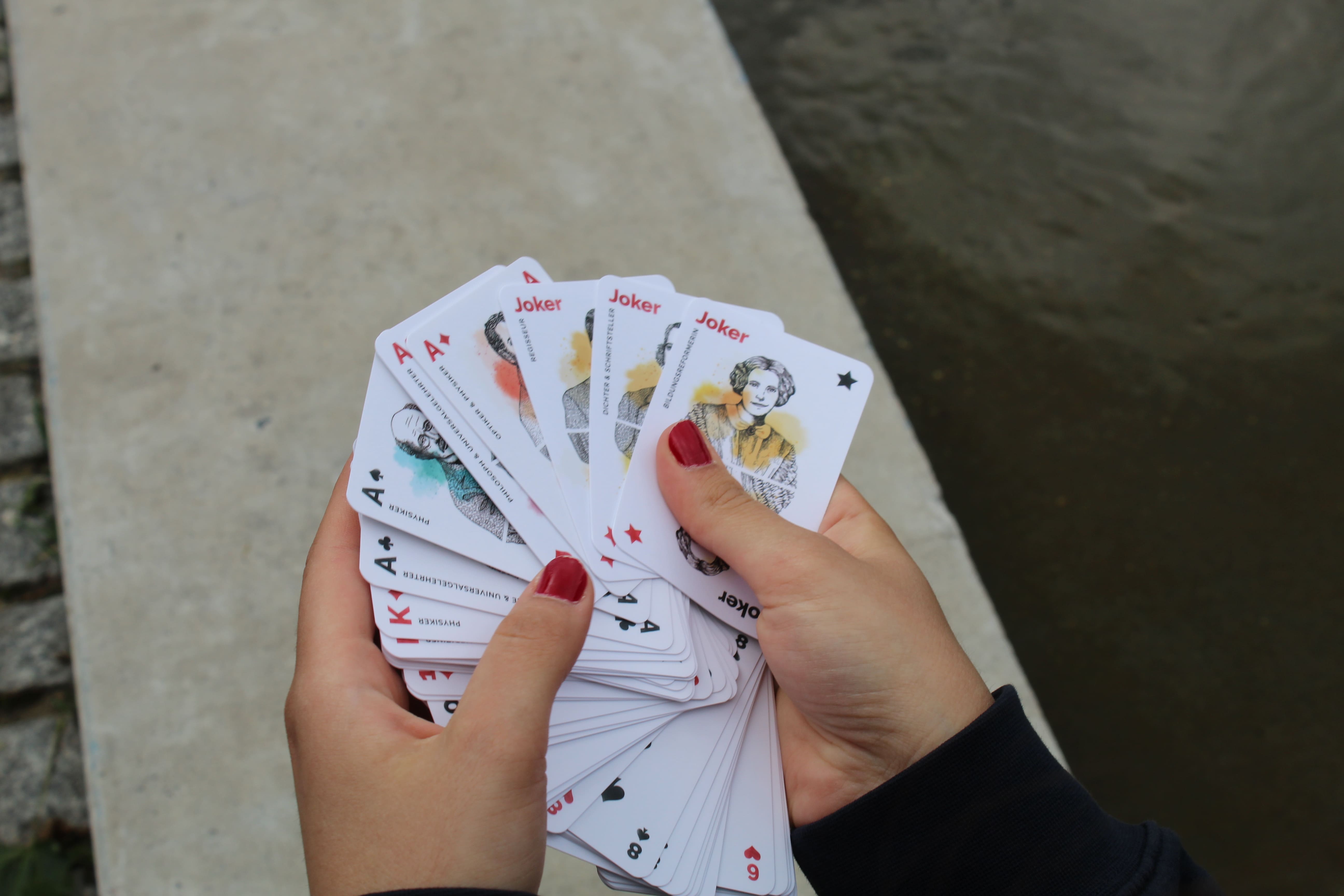 Skat und Romme. Abgebildet sind zwei Hände, die ein buntes Kartenspiel aufgefächert in der Hand tragen.