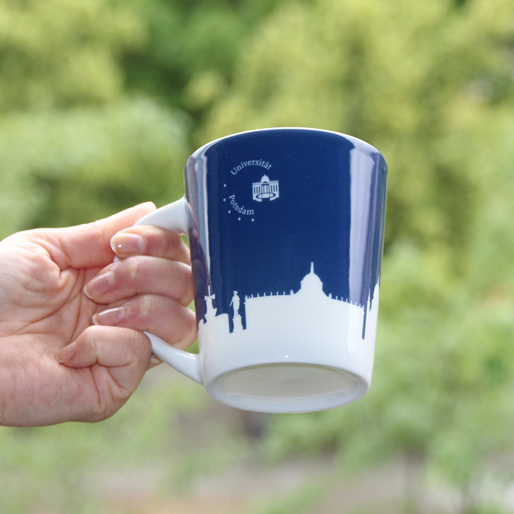 Porzellan-pot. Abgebildet ist eine blaue Tasse mit einer weißen Silhouette der Stadt Potsdam und zusätzlich dem Universitätslogo. Im Hintergrund erkennt man verschwommen ein paar Bäume.