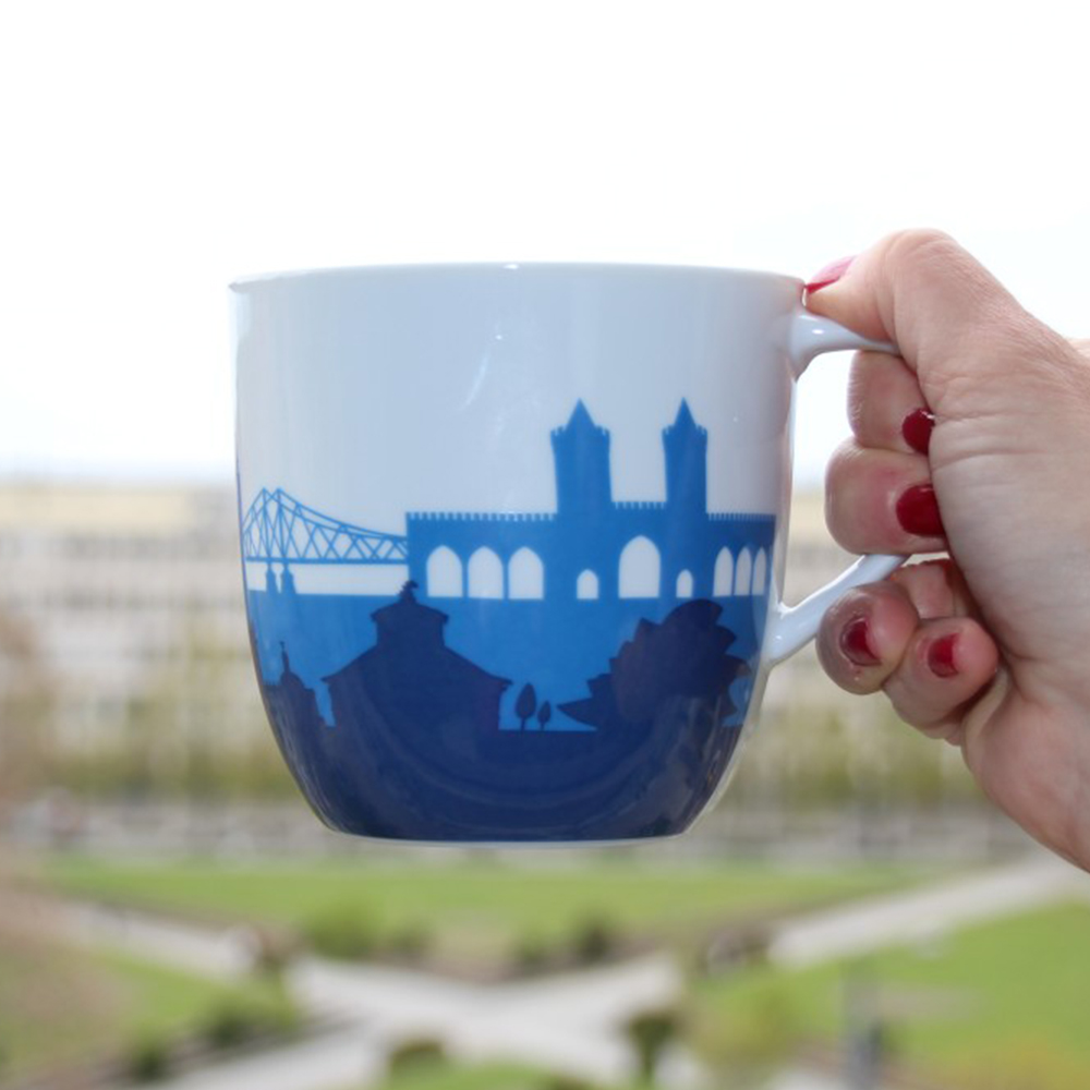 Jumbo-tasse. Abgebildet ist eine große weiße Tasse mit einem blauen Brückenlogo. Im Hintergrund sieht man Wiesen und Wege.