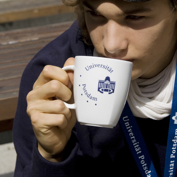 Tasse weiß. Ein junger Mann trinkt aus der weißen Universitätstasse. Er sitzt im Freien auf einer Bank.