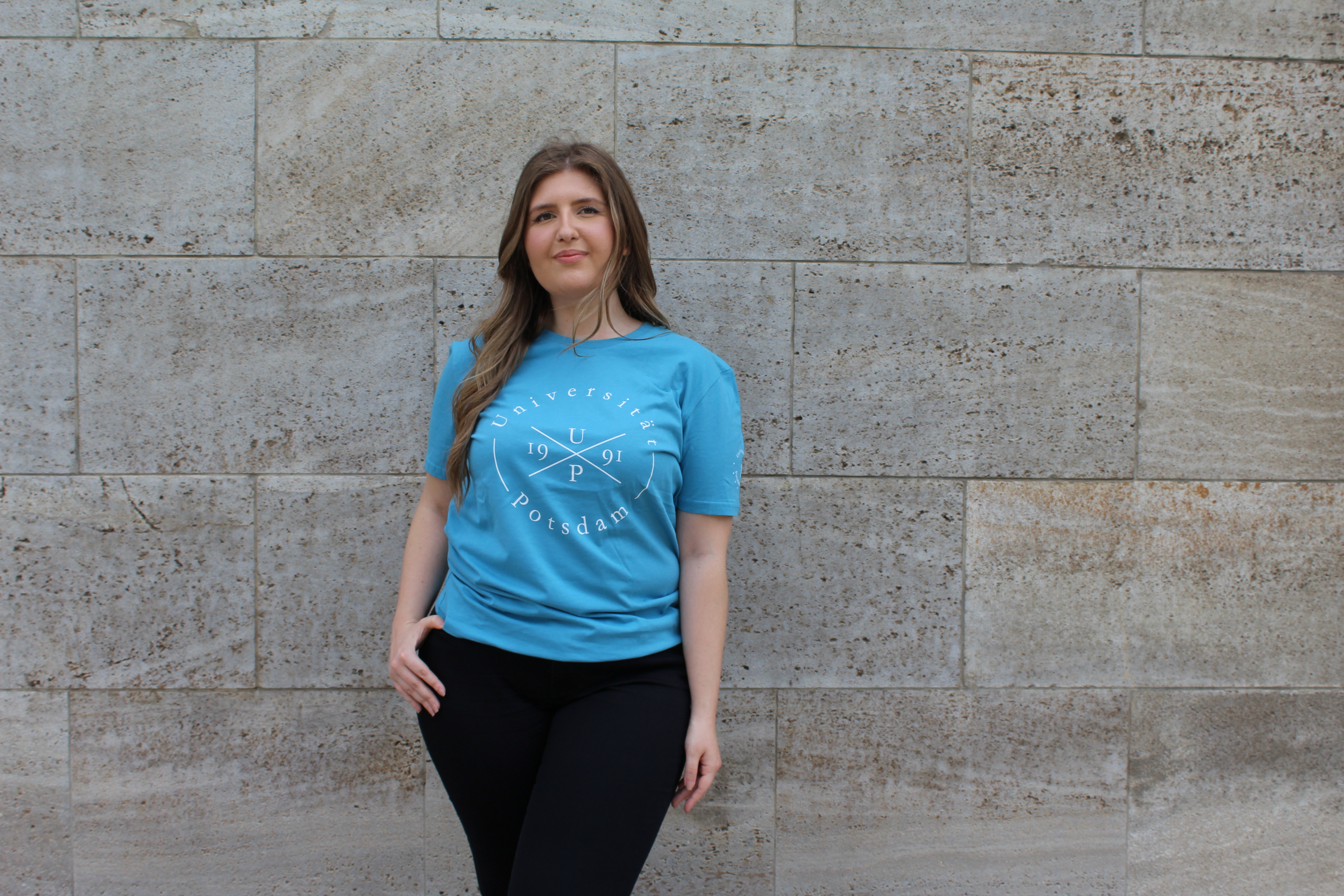 Styler T-shirt. Abgebildet ist eine junge Frau, die vor einer Wand posiert. Sie trägt ein blaues Styler T-shirt.