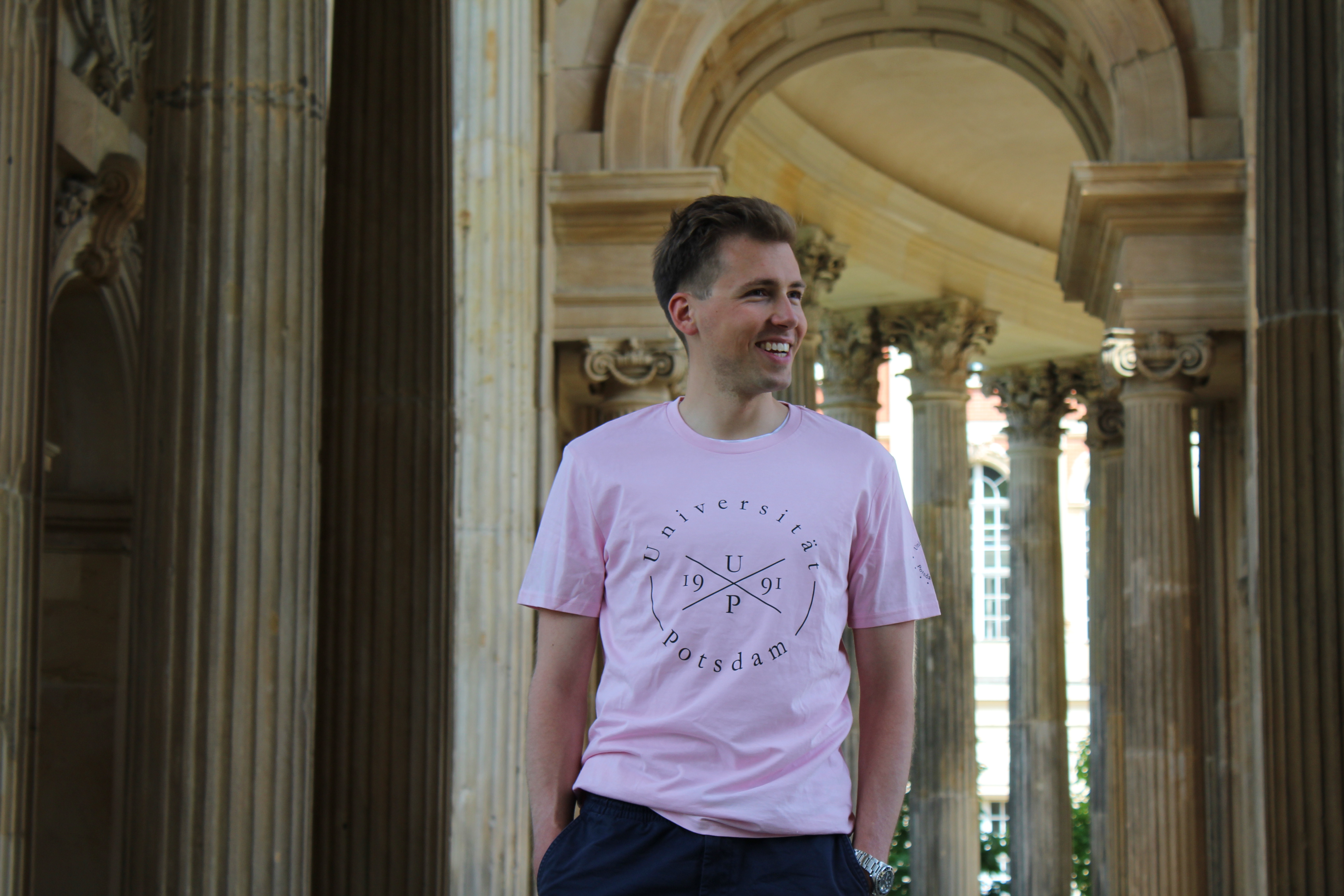 Tshirt- Styler. Abgebildet ist ein  junger Mann mit einem rosa farbenen Styler  T-shirt.  Im Hintergrund erkennt man  Säulen.