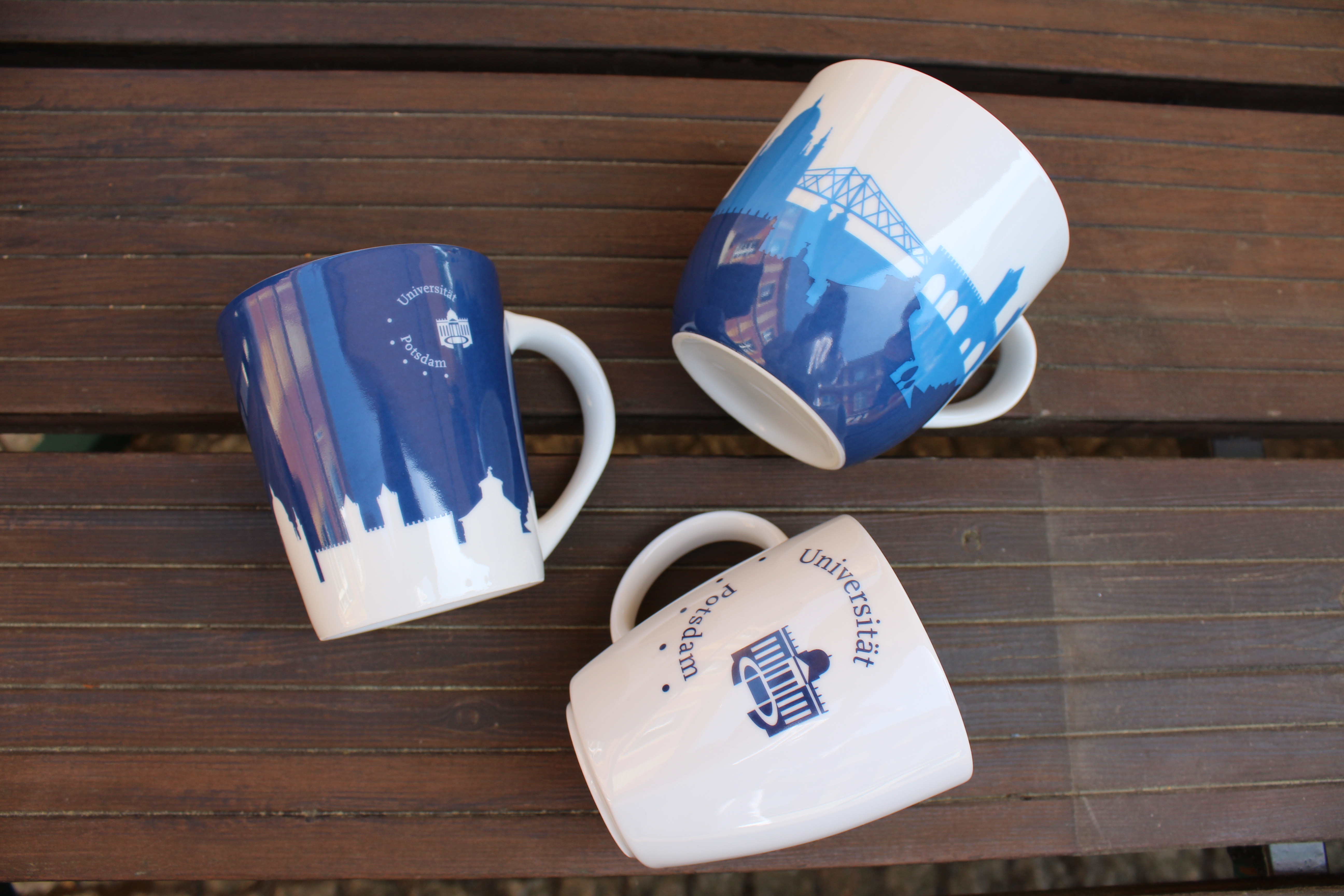 Jumbo Tasse, Tasse weiß, Pott blau. Abgebildet sind die 3 verschiedenen Tassen des Unishop Potsdams auf einer Holzunterlage.