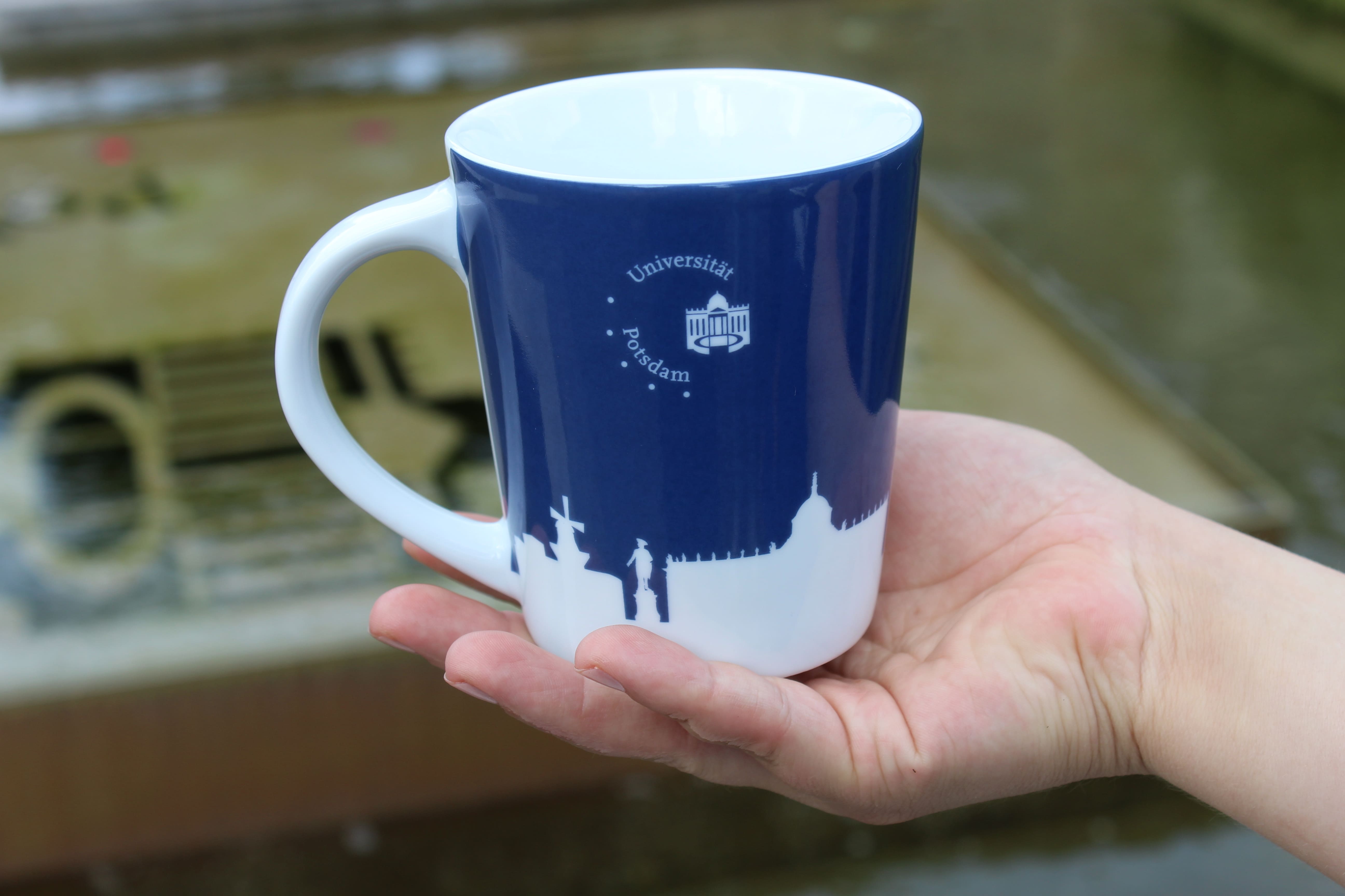 Porzellan-pot. Abgebildet ist eine blaue Tasse mit einer weißen Silhouette der Stadt Potsdam und zusätzlich dem Universitätslogo. Im Hintergrund erkennt man verschwommen ein Gebäude.