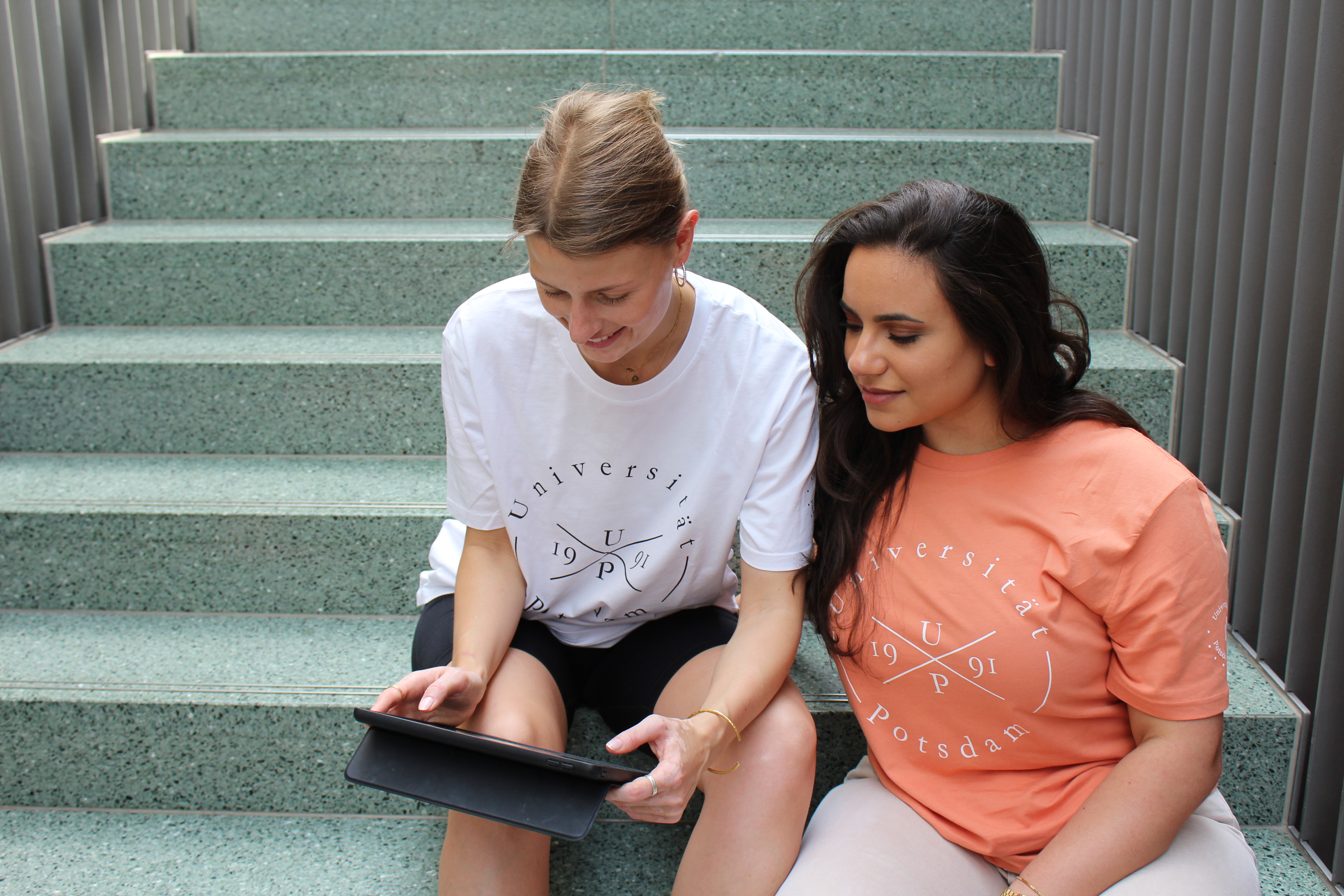 Styler T-shirts. Abgebildet sind zwei junge Frauen, die auf einer Treppe sitzen. Die Frau links trägt ein weißes Styler T-shirt. Die Frau rechts von ihr ein orange farbenes Styler T-shirt.